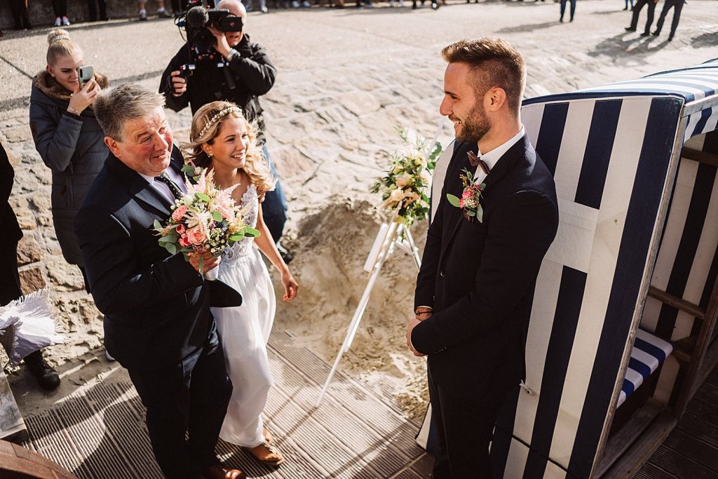 Braut wird von ihrem Vater zum Badekarren geführt, der Bräutigam wartet lächelnd am Strand von Norderney.