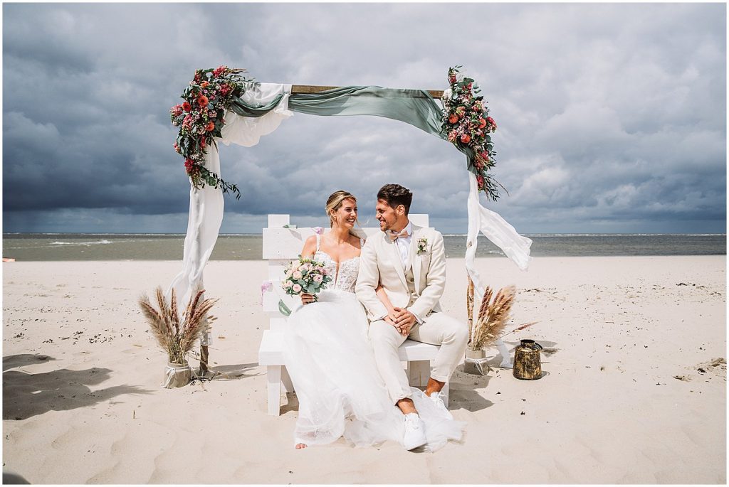 Braut und Bräutigam sitzen entspannt nach der freien Trauung unter dem Traubogen am Strand von Baltrum – natürliche Hochzeitsreportage mit Meerblick und Nordseeluft