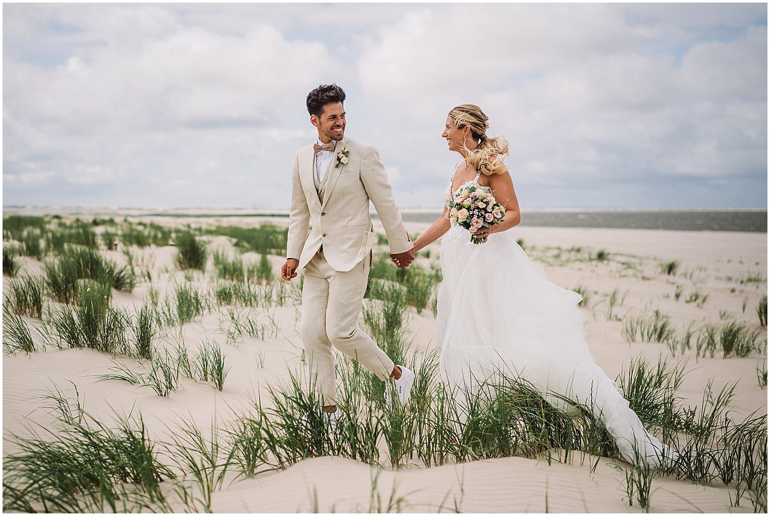 Braut und Bräutigam laufen Hand in Hand durch die Dünen auf Baltrum – natürliche Hochzeitsreportage am Strand der ostfriesischen Insel mit Wind, Weite und Nordseelicht