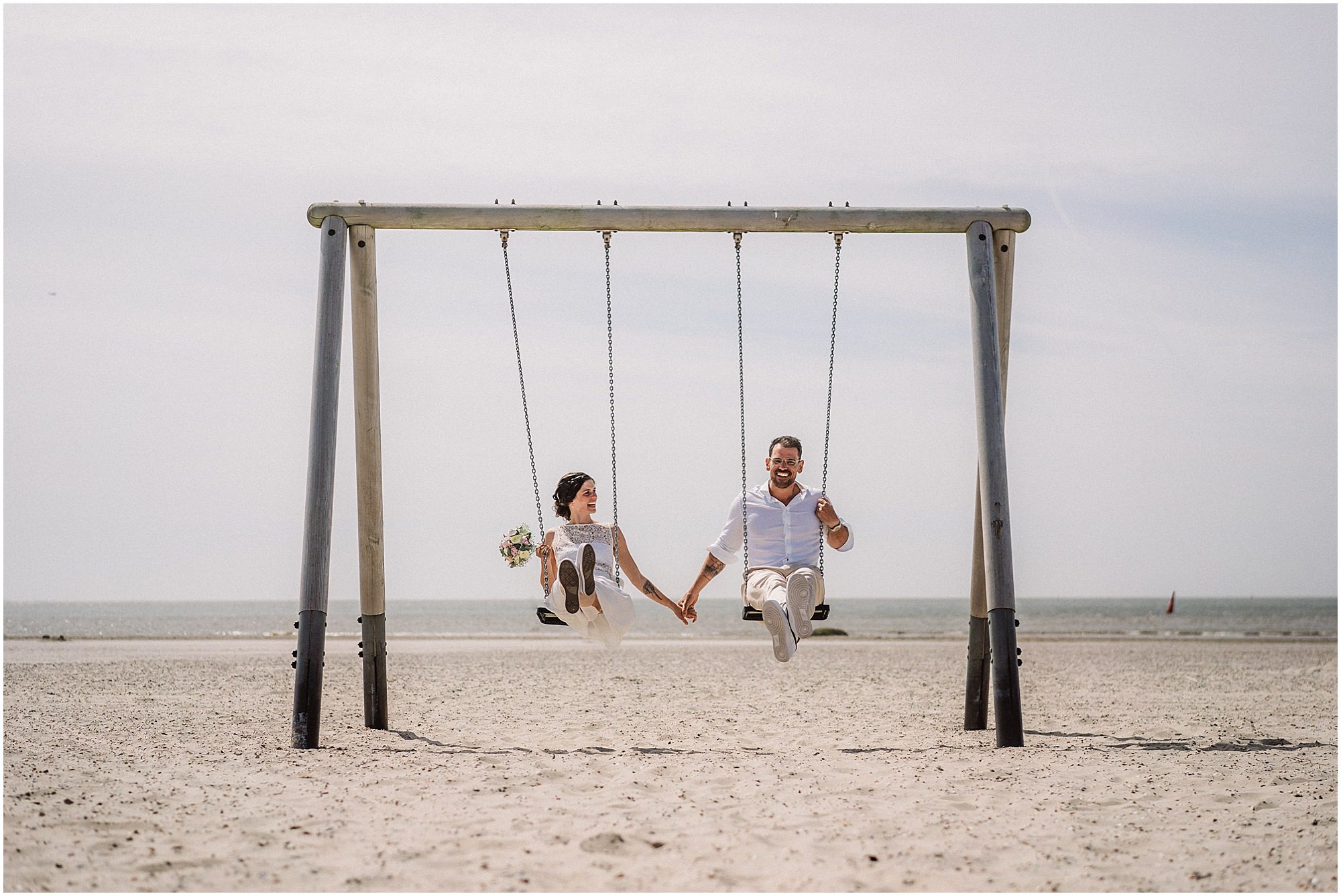 Braut und Bräutigam lachen am auf einer Schaukel am Strand bei einer Inselhochzeit auf Norderney